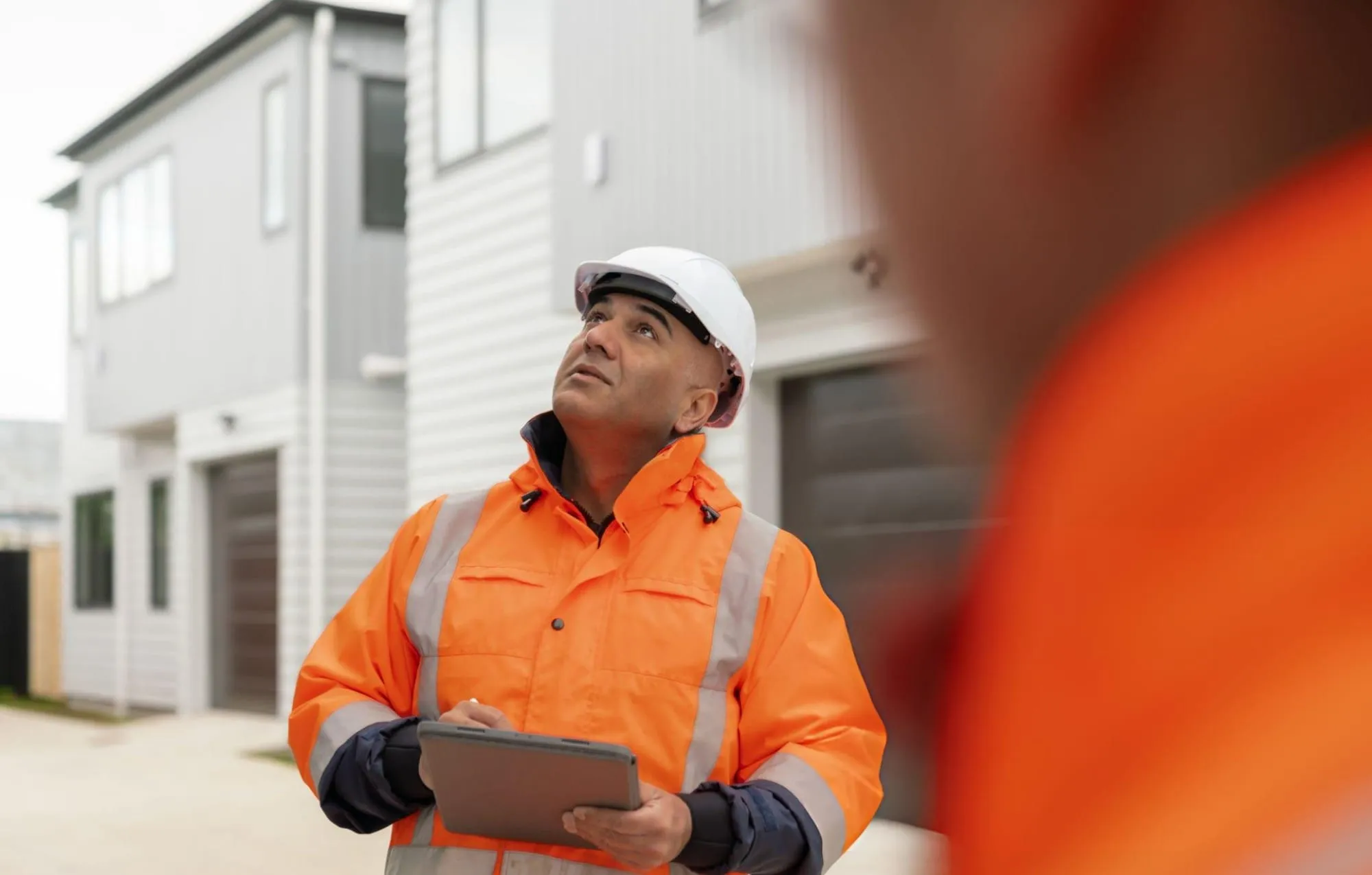 Construction supervisor in a hi-vis jacket and white hard hat holds a tablet while looking up at new townhomes