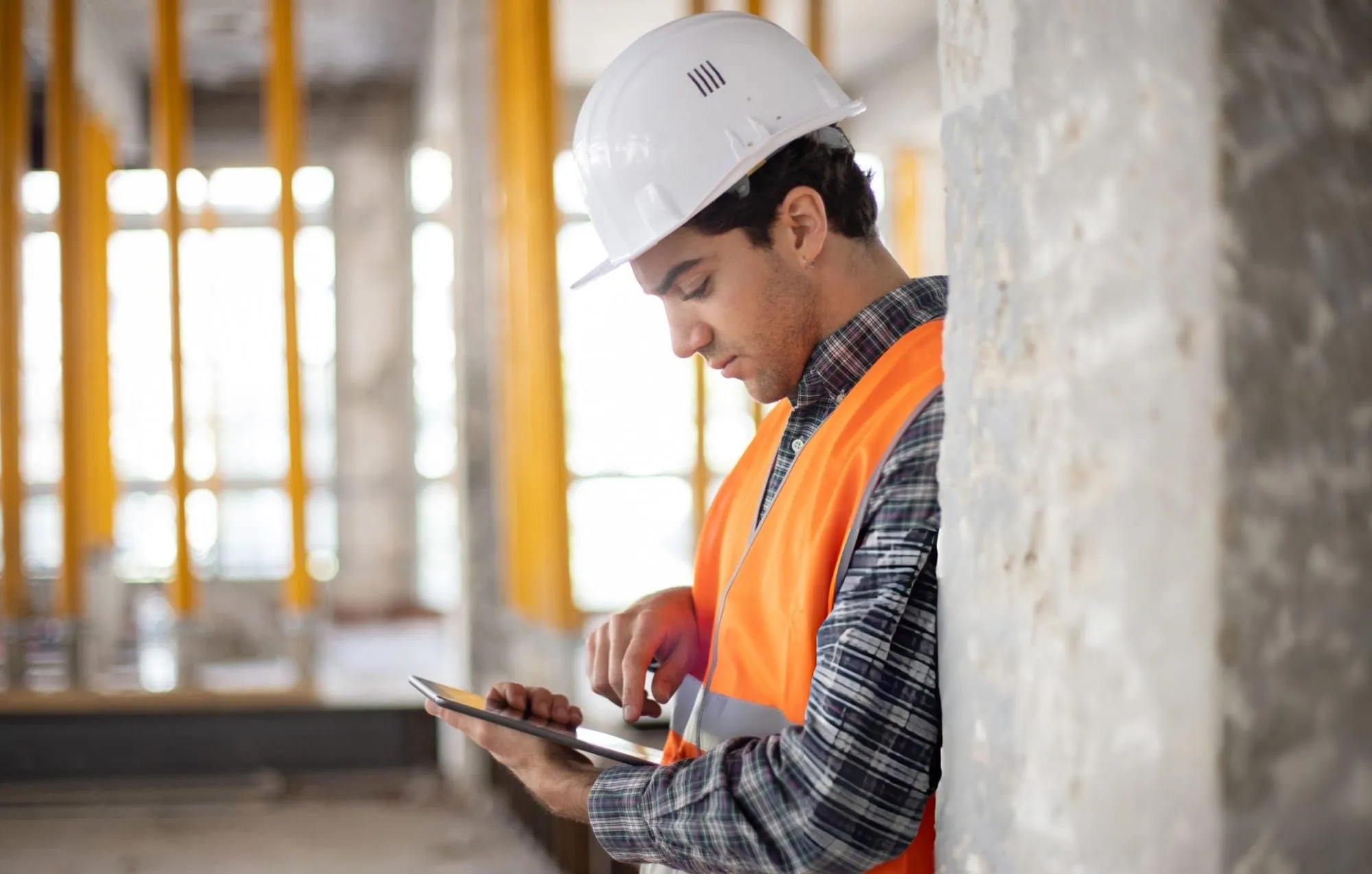 Construction worker in a white hard hat and orange vest using a tablet inside an unfinished building