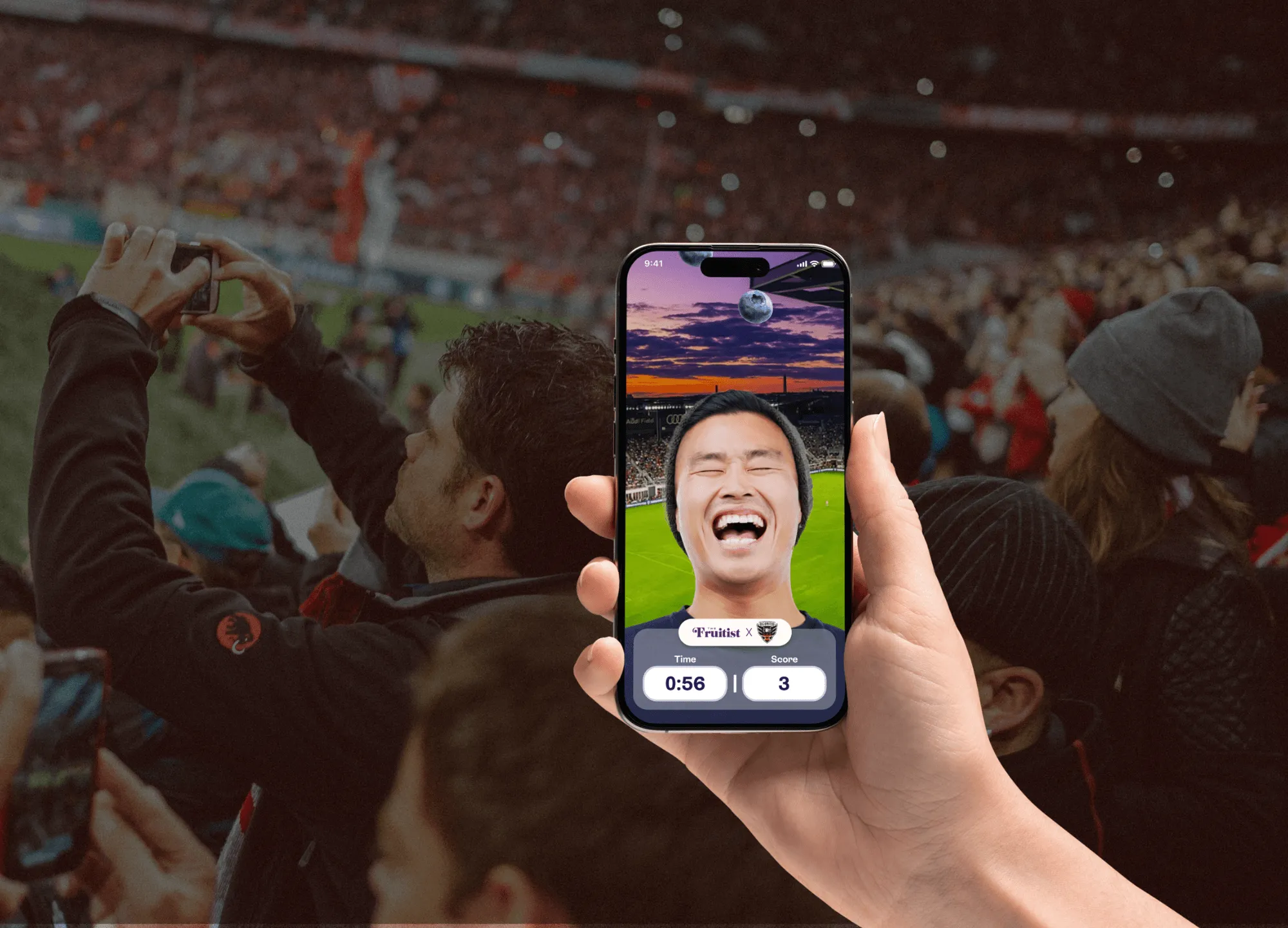 Hand holding a phone showing the Blueberry Catch AR game with a laughing fan, in front of a stadium crowd
