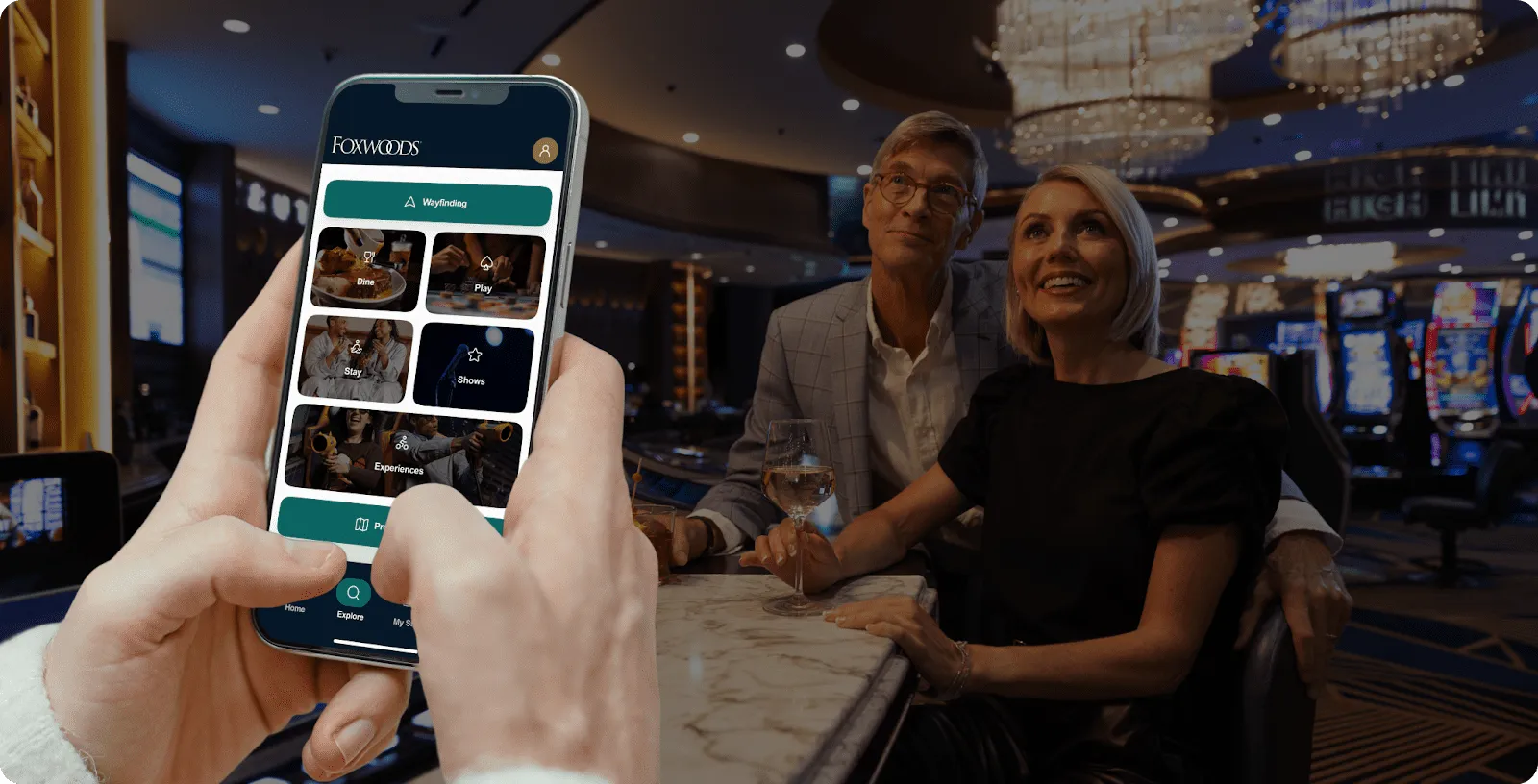 Hand holding a phone showing the Foxwoods app Wayfinding button and tiles for dining, play, stay, shows, and experiences