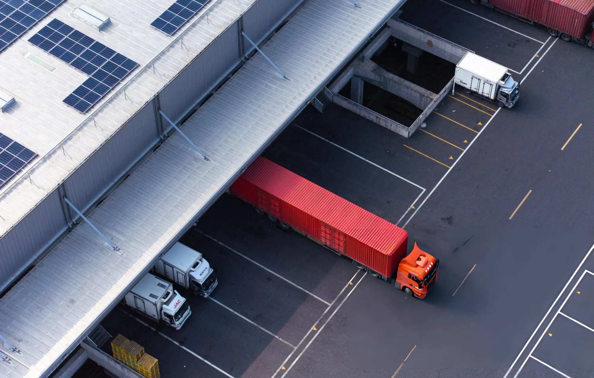Aerial view of a red cargo truck backed up to a warehouse loading dock beside delivery trucks, under a solar-panel roof