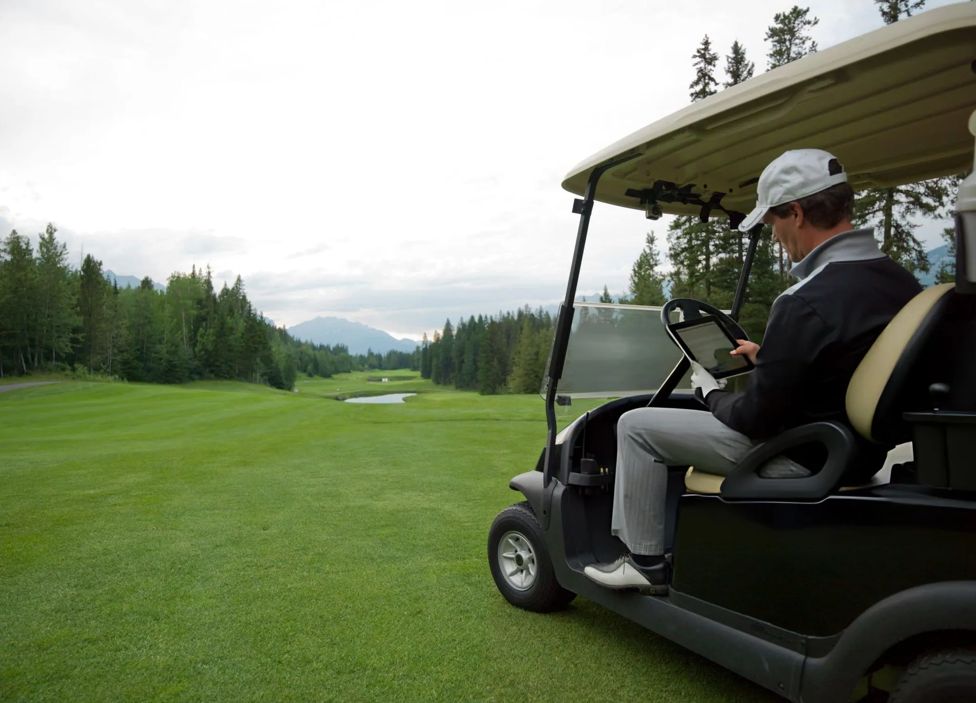 Golfer in a cap and glove checking a tablet from a golf cart parked on a fairway with forested hills