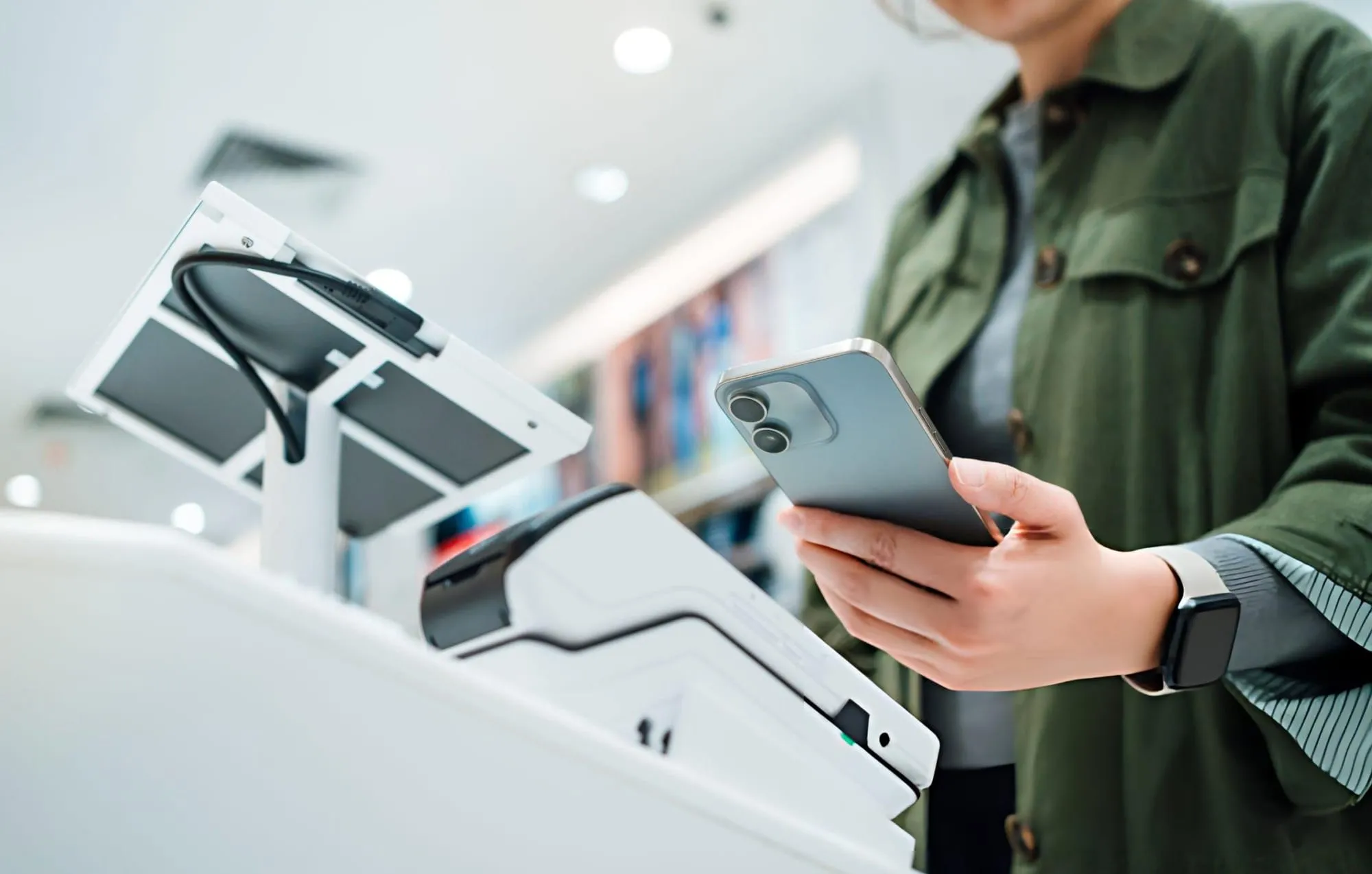 Shopper in a green jacket holding a phone near a card reader at checkout