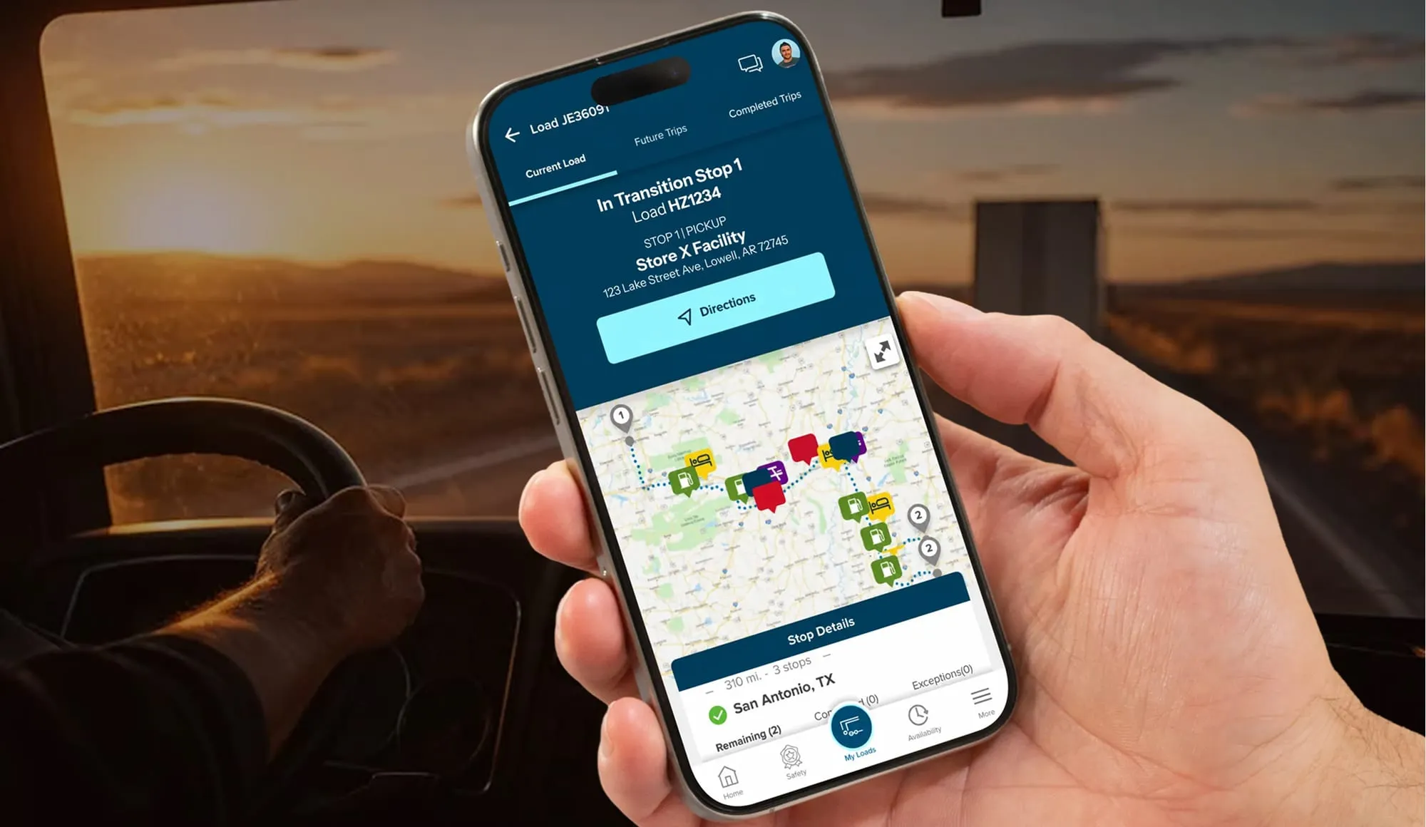 A driver's hand holding a phone with the driver app open to a load in transit, with a truck cab and a sunset road behind.