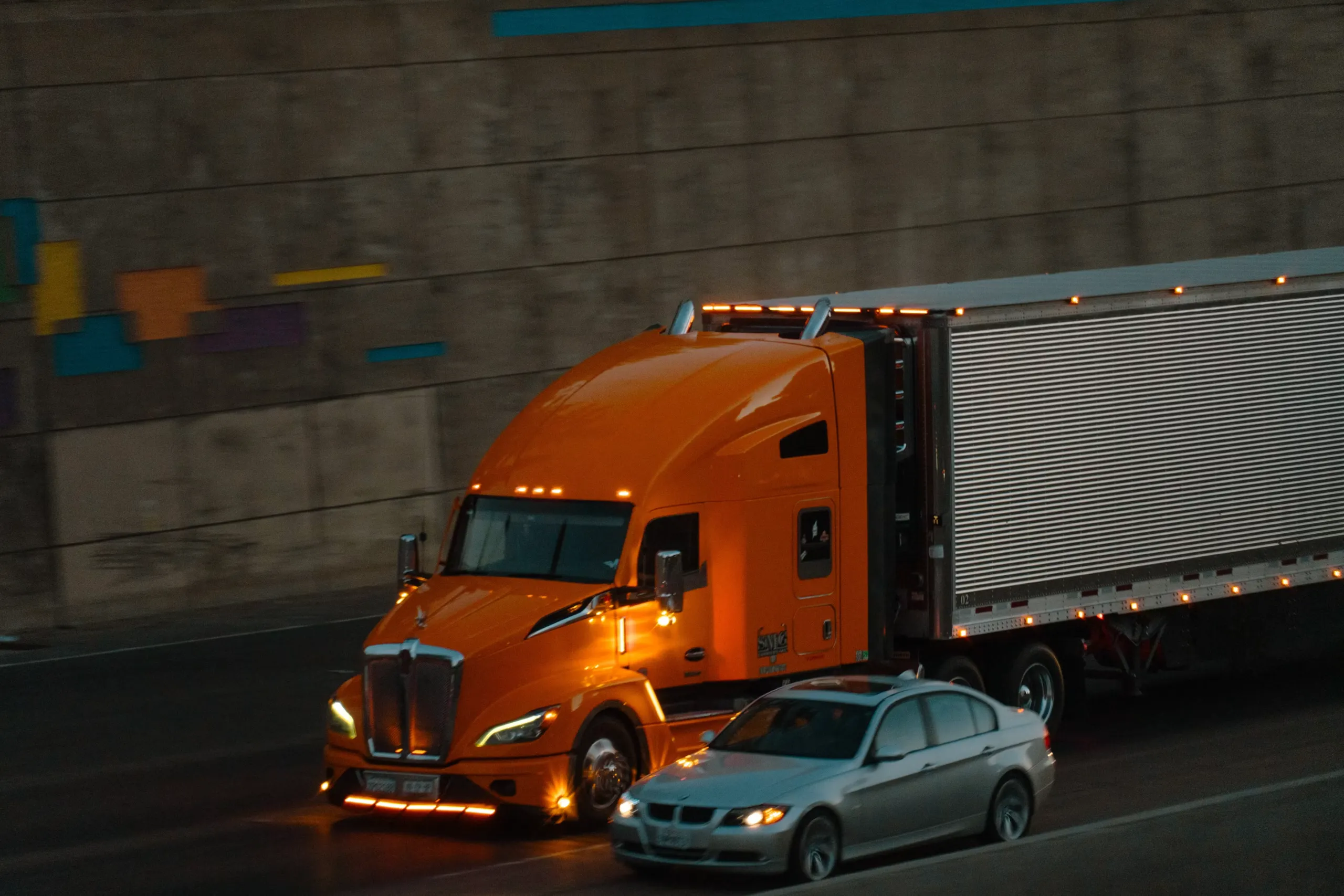 Orange semi truck pulling a trailer at night beside a sedan on a highway