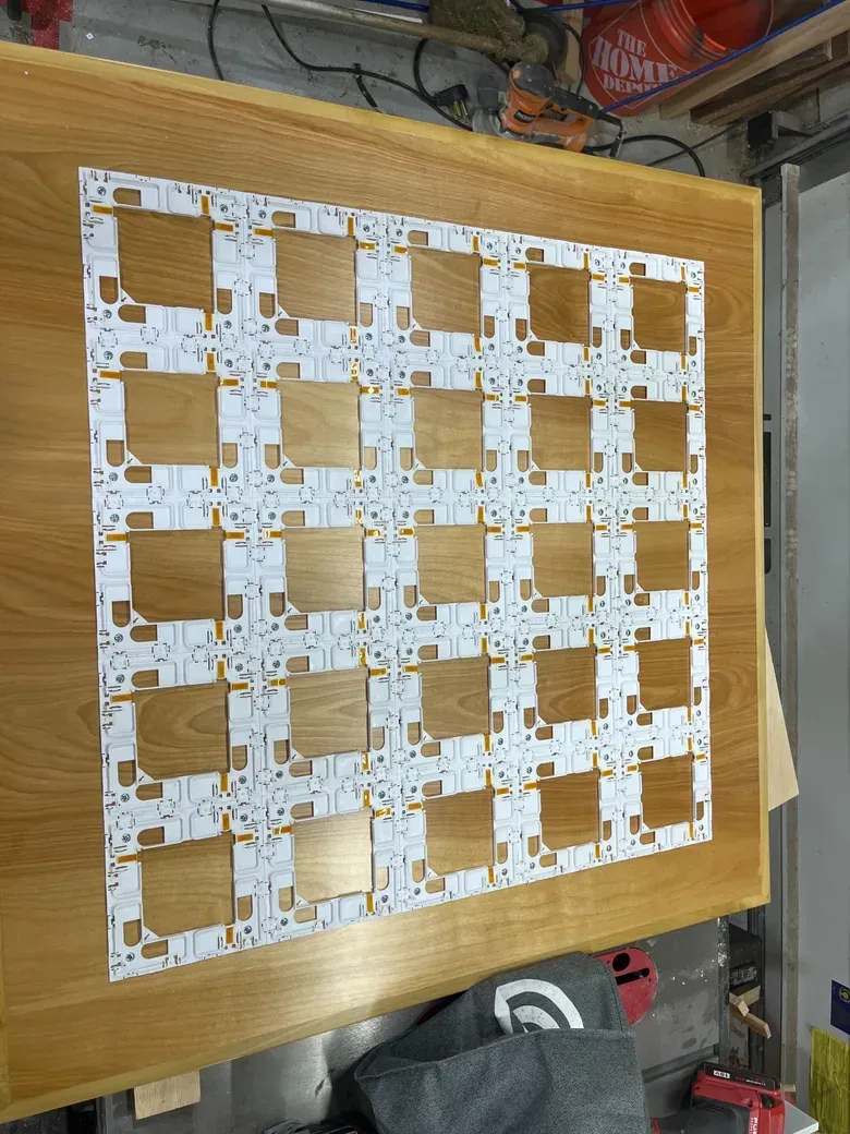 A five by five grid of white circuit-board light panels laid flat on a wooden tabletop in a workshop, before they were mounted.