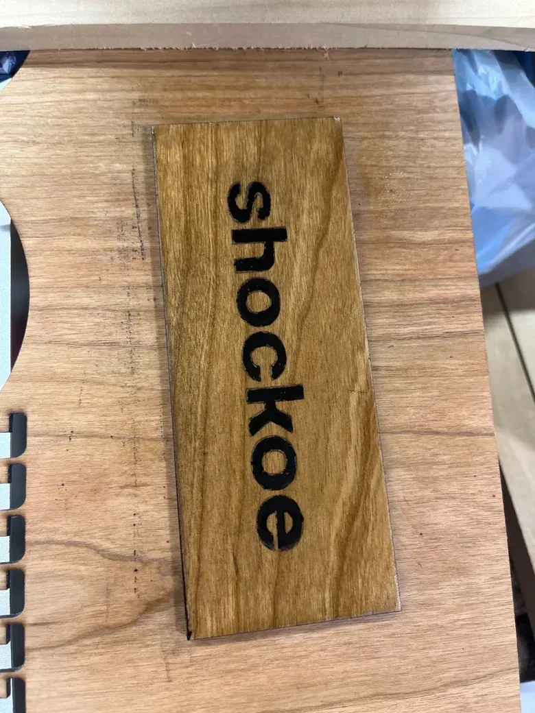 A wooden nameplate with a dark engraved wordmark, set on a piece of raw wood before it was mounted.