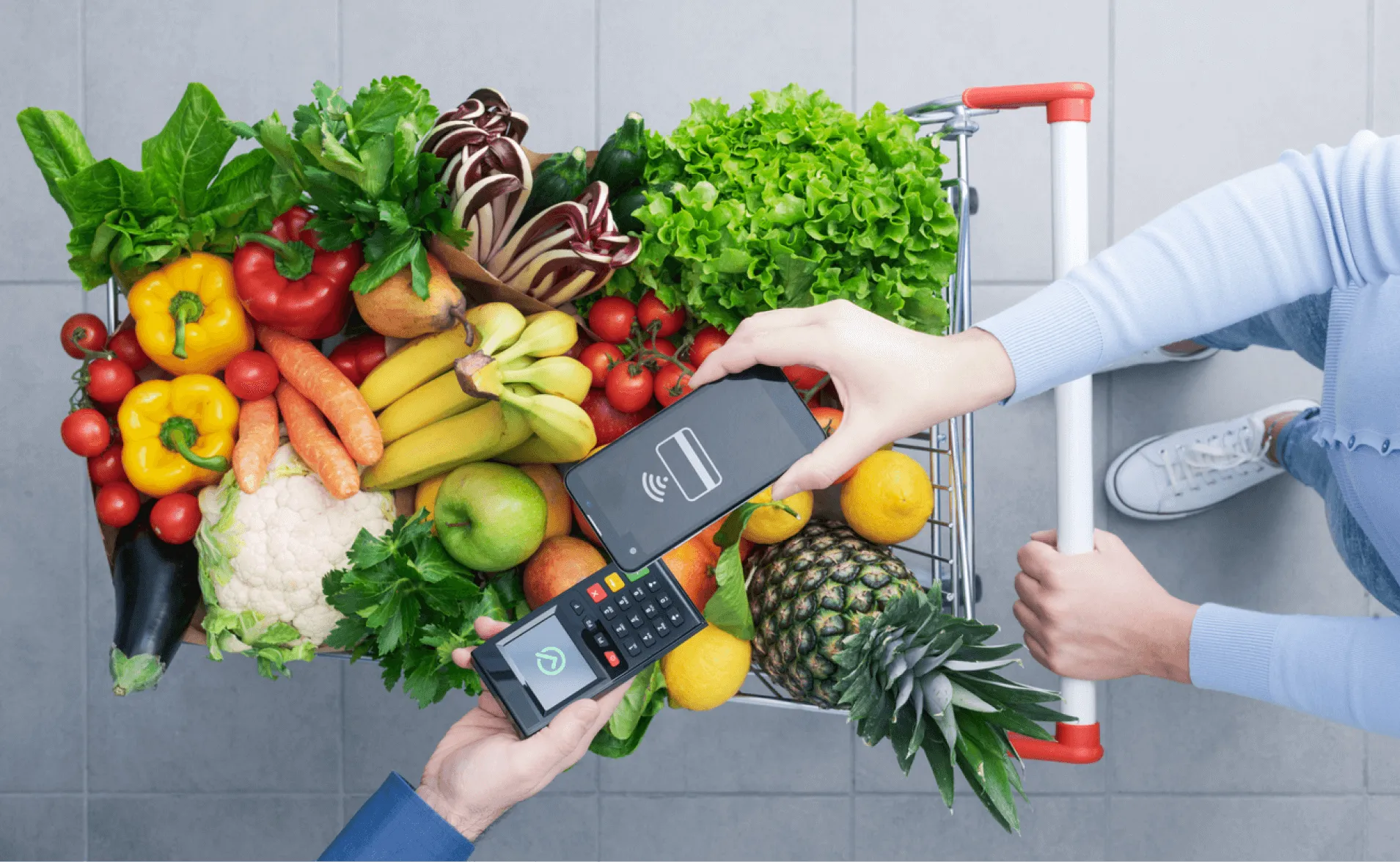 Overhead view of a shopper paying with a phone at a handheld card reader over a cart of fresh produce