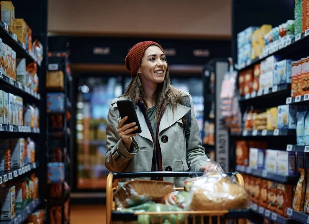 Smiling shopper in a red knit hat checking her phone in a grocery aisle while pushing a cart