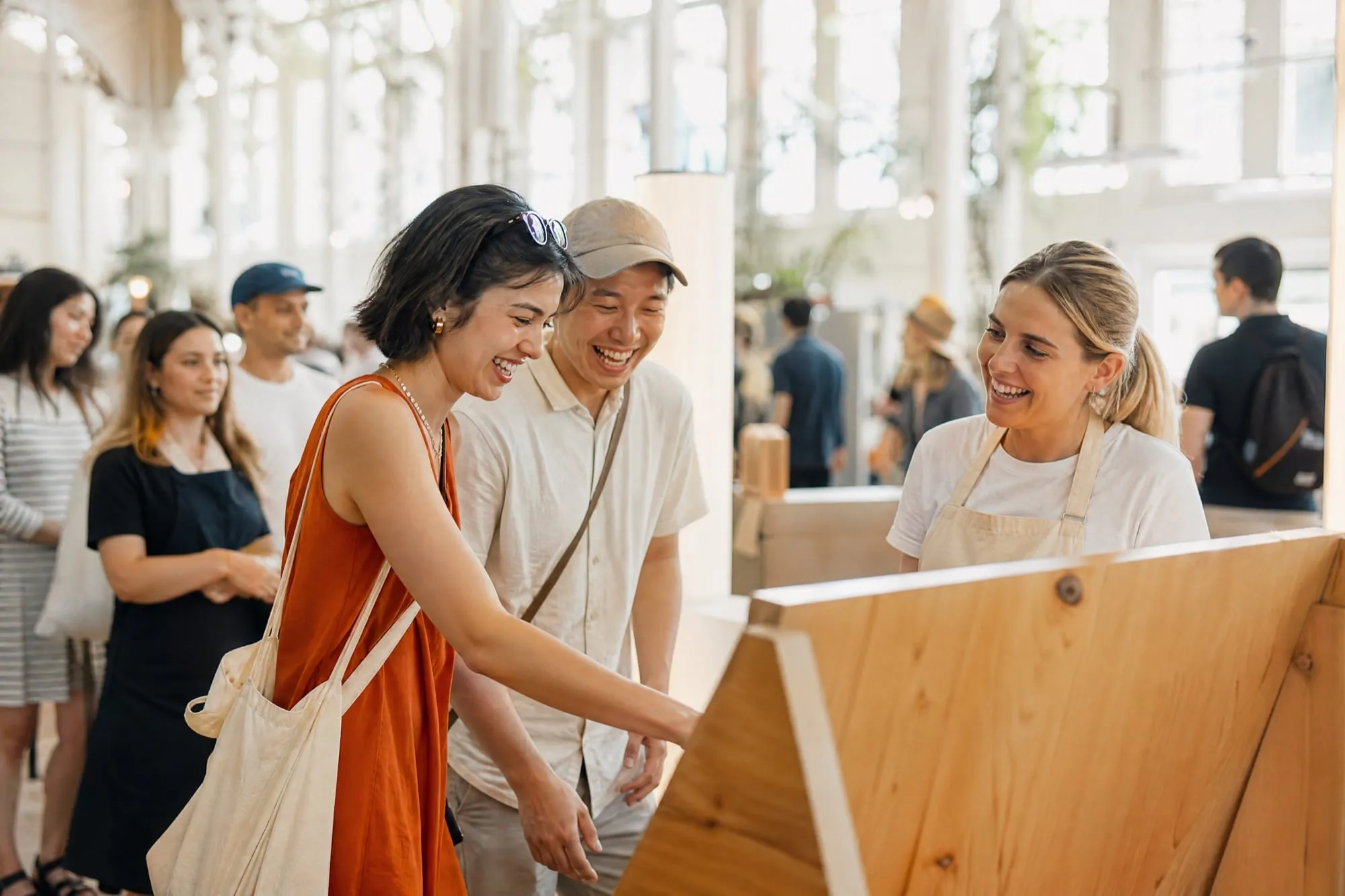 Two shoppers laughing as they try a wooden interactive display while a brand host in a cream apron watches