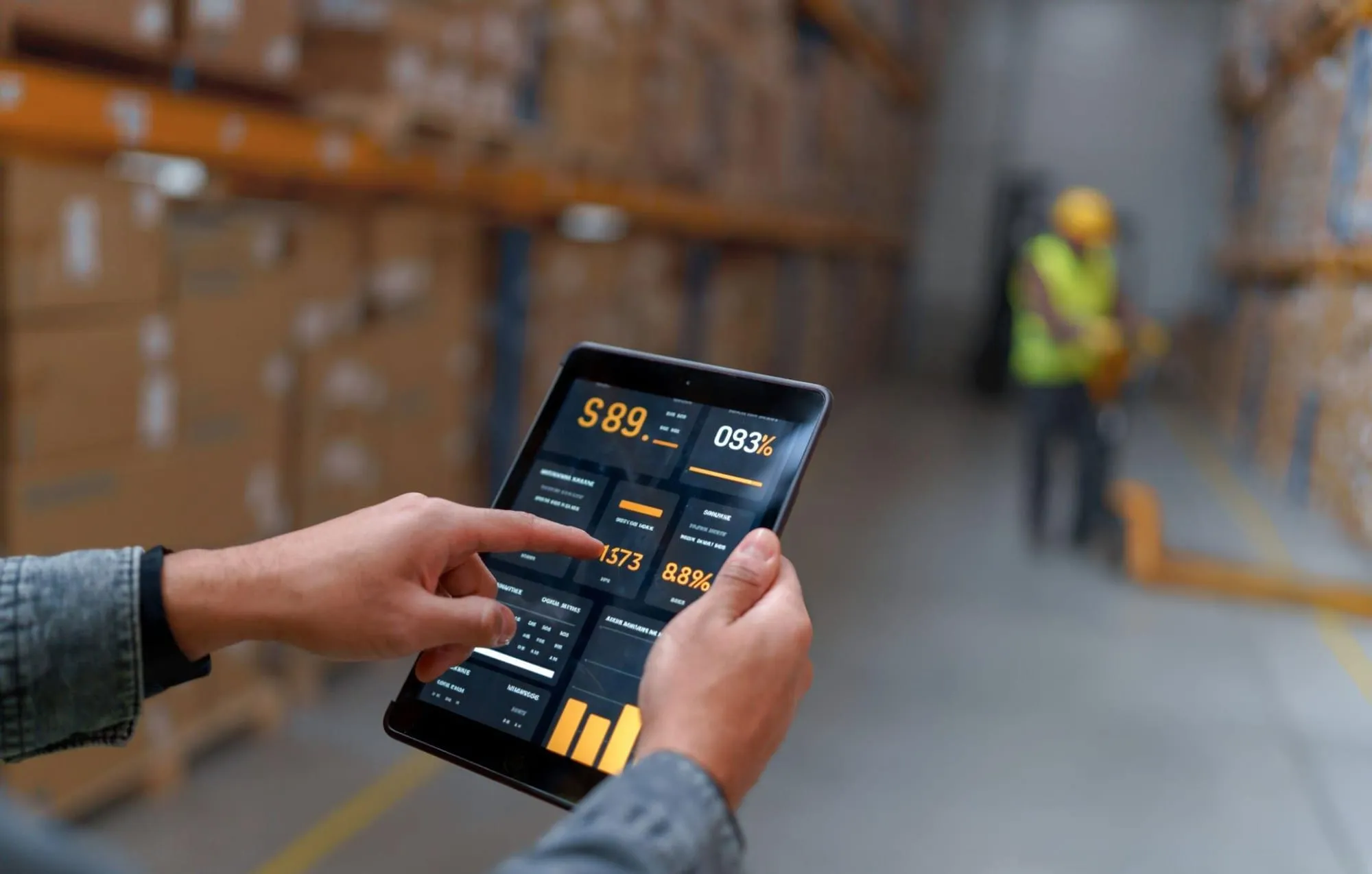 Hand tapping a tablet dashboard of logistics figures in a blurred warehouse