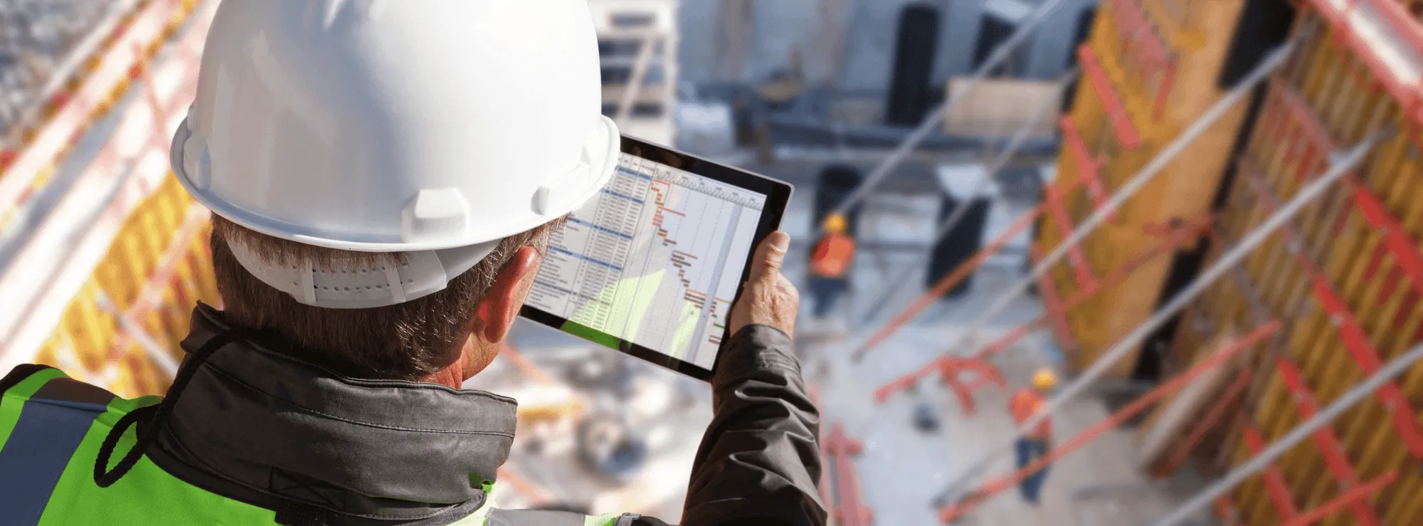 Builder in a white hard hat viewing a project schedule on a tablet above a construction site with formwork